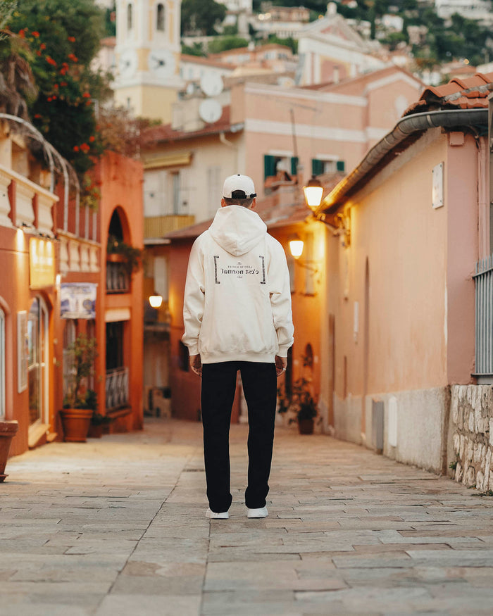 A person viewed from the back wearing Le Hoodie in cream, featuring the FRC mirror logo, standing in a narrow street in the French Riviera. They are also wearing black pants and a white c'est normal cap.