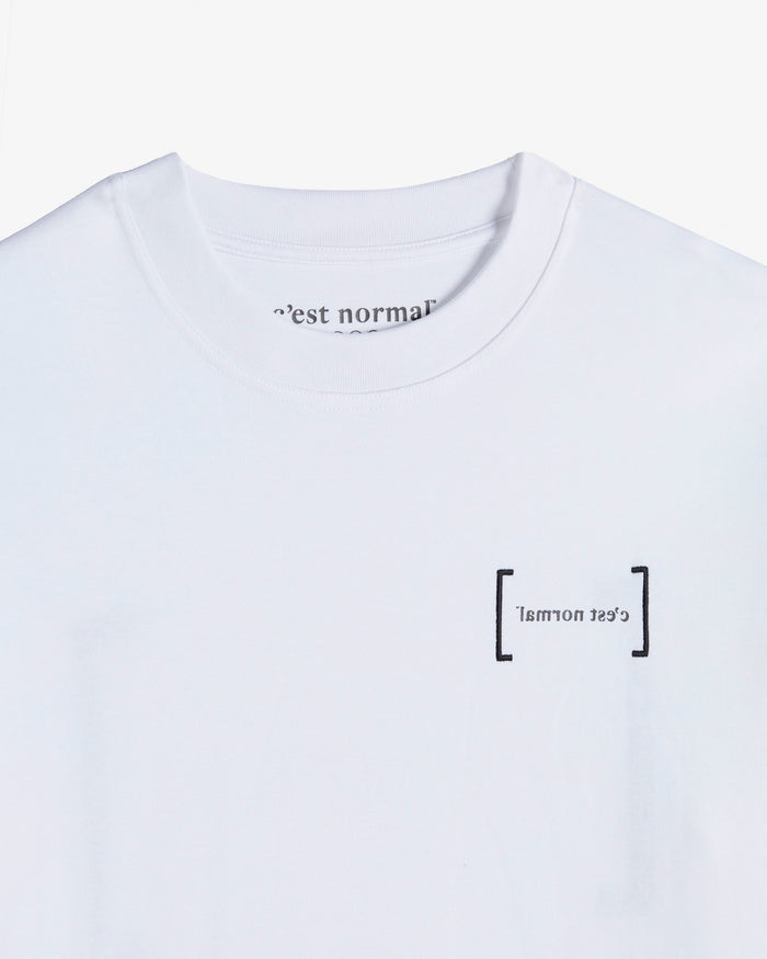 A close-up shot of the white Le T-Shirt, highlighting the classic ribbed collar and the black FRC Edition mirror logo on the chest. The inside collar reveals the c'est normal branding.