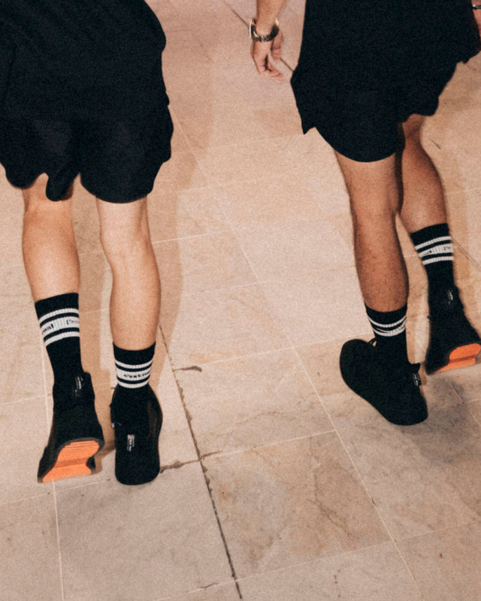 A back view of two people walking, wearing Les Socks in black. The image highlights the white stripes and the c'est normal logo at mid-calf, paired with black shorts and black sneakers.