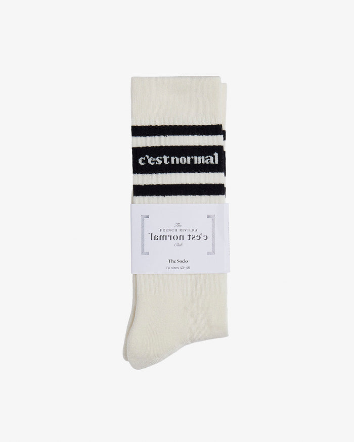 A folded pair of Les Socks in cream, laid flat on a white background. The image highlights the black stripes, the knitted c'est normal logo, and the French Riviera Club paper wrapper.