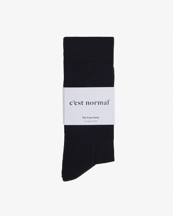 A pair of black The Athlétique Socks laid flat on a white background, folded and wrapped in a white paper sleeve featuring the c'est normal logo.