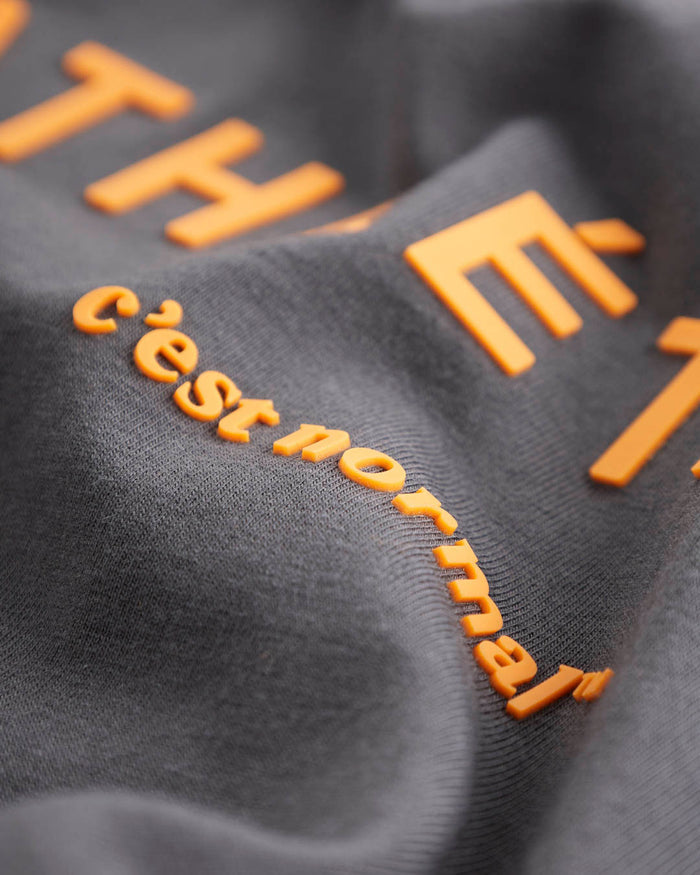 A close-up shot of the asphalt fabric of The Athlétique Tee, highlighting the raised orange Athlétique and c'est normal logos.