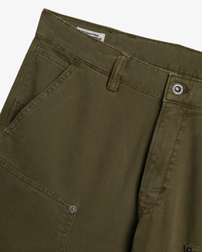 A close-up shot of the front of The Carpenter Pant in olive green, showcasing the textured fabric, a slash pocket, and custom c'est normal buttons and rivets.