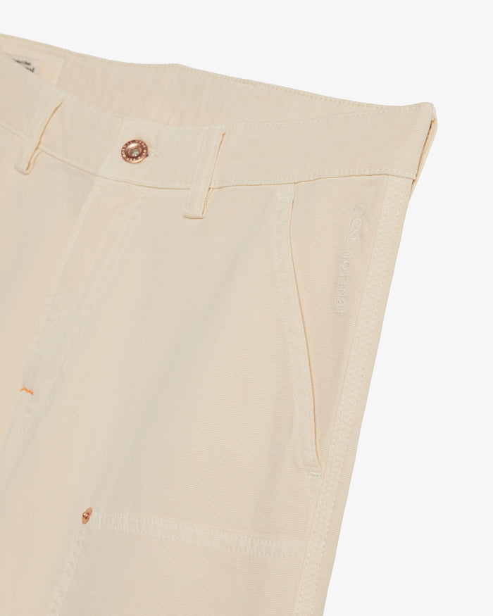 A close-up view of The Carpenter Pant in cream on a white background, focusing on the front slash pocket, copper-toned rivet, and the subtle embroidered c'est normal logo along the side seam.