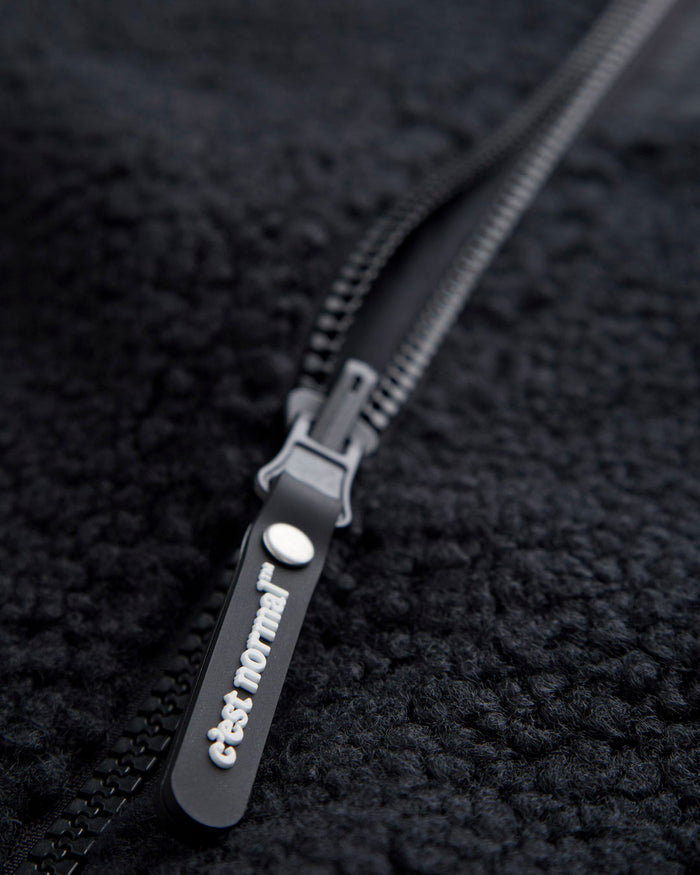 A close-up shot of the black pile fleece material of The Reversible Pile Jacket, focusing on the tone on tone zipper and the black rubber puller with the white c'est normal logo.