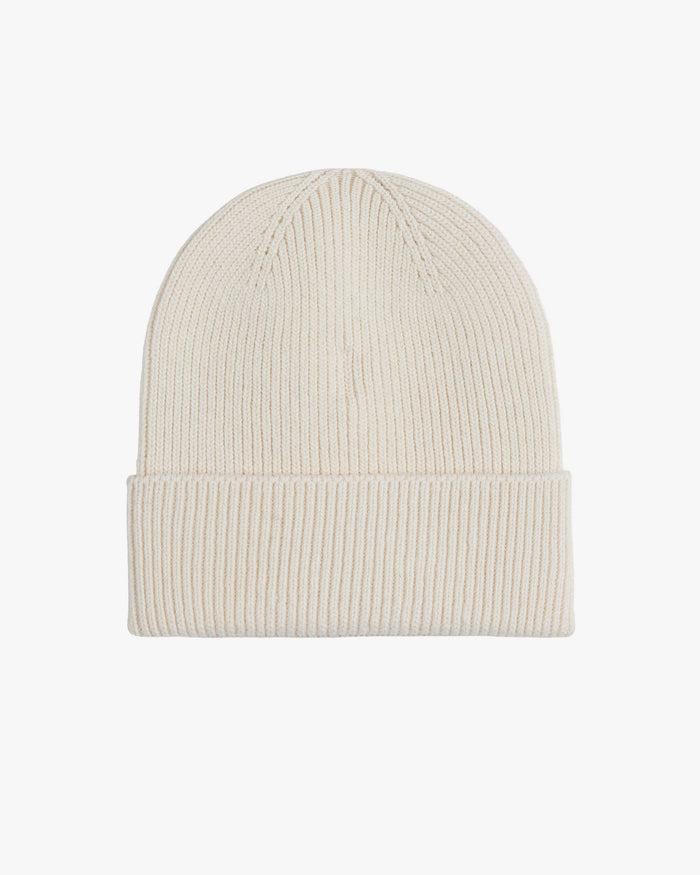 The Ribbed Beanie in cream, laid flat on a white background, highlighting its classic ribbed knit texture and folded brim. The tone on tone rubber tag with the c'est normal logo is not visible from this angle.