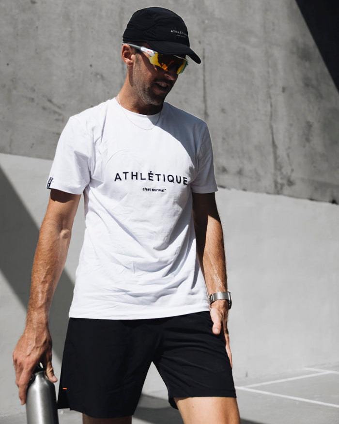 A man wearing The Running Cap in black, sunglasses, and a white t-shirt, standing outdoors in front of a concrete wall.