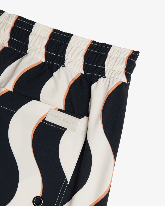 A detailed close-up of The Shorts in Noir Curl Stripe, focusing on the elasticated waistband and the pocket with the c'est normal logo patch. The image also showcases the wavy black, white, and orange striped pattern.