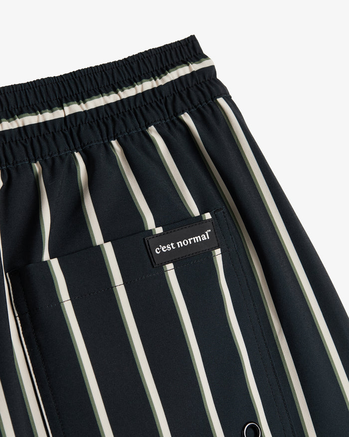 A detailed close-up of The Shorts in Noir Stripe, focusing on the elasticated waistband and the back pocket with a black patch featuring the white c'est normal logo. The image also showcases the striped pattern in black, white, and green.