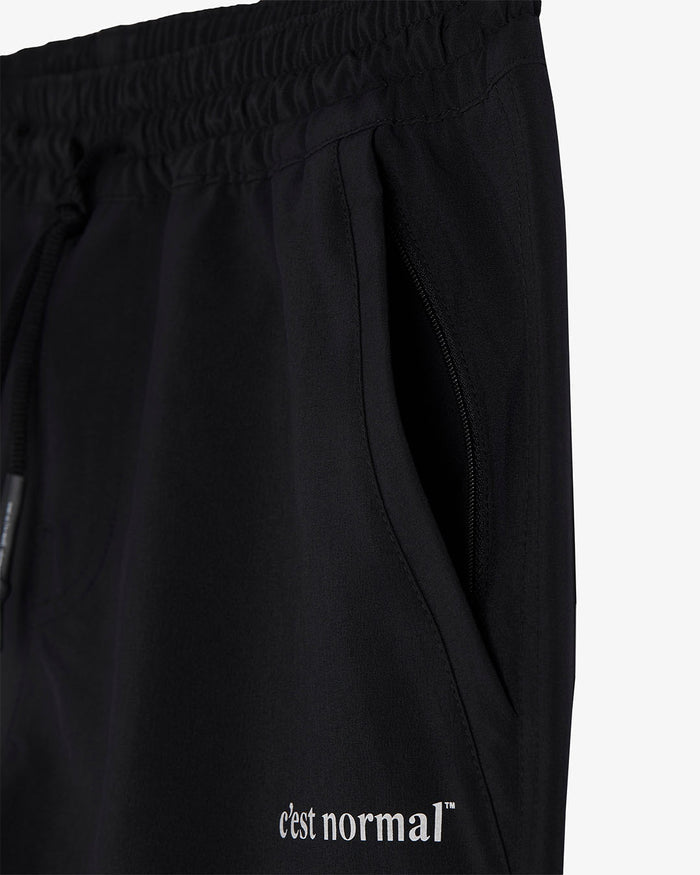 A detailed close-up of The Shorts in black, highlighting the zippered side pocket, elastic waistband with drawstrings, and the white c'est normal logo on the lower leg.