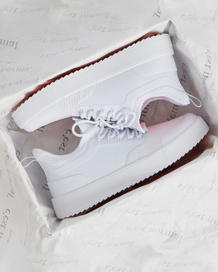 A pair of The Sneakers 2.0 in white, resting inside a shoebox lined with c'est normal branded tissue paper. The image highlights the tone on tone mesh upper, custom lace loops, and thick white rubber soles.