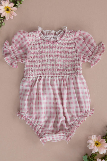 The Ivy City Co Baby Cordelia Romper in Desert Rose Gingham, featuring a smocked bodice, puffed short sleeves, ruffled collar, and snap buttons at the bottom, is displayed on a beige background with pink flowers in the corners.