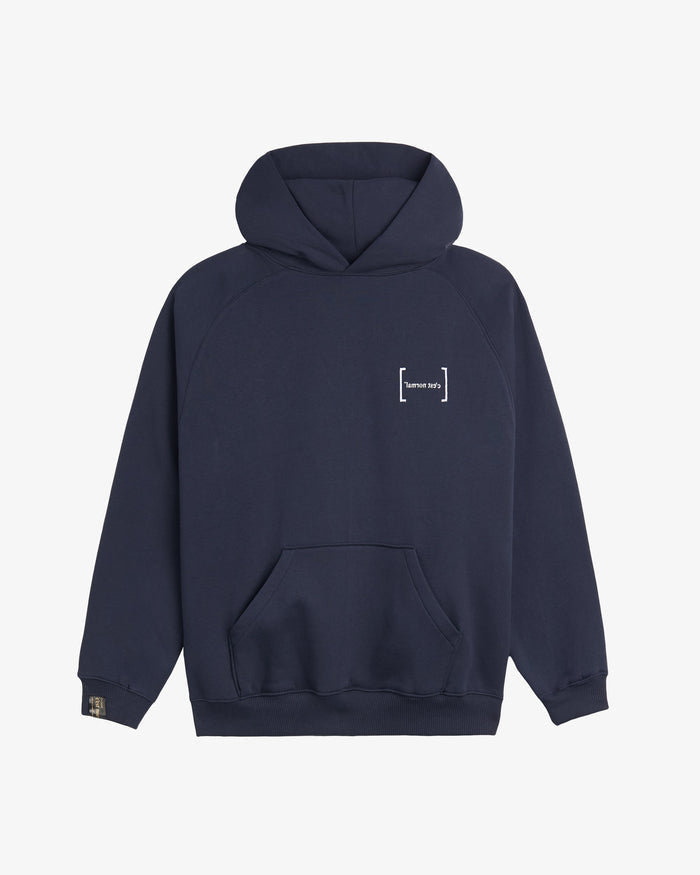 A front view of Le Hoodie in navy, laid flat on a white background. The image highlights the kangaroo front pocket, the white FRC mirror c'est normal logo on the chest, and the signature cuff tag on the sleeve.