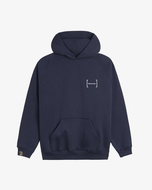 A front view of Le Hoodie in navy, laid flat on a white background. The image highlights the kangaroo front pocket, the white FRC mirror c'est normal logo on the chest, and the signature cuff tag on the sleeve.