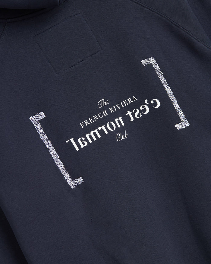 A detailed close-up of Le Hoodie in Navy, showcasing the center back featuring the white FRC mirror logo, which includes the text "The FRENCH RIVIERA Club" and a mirrored c'est normal logo.