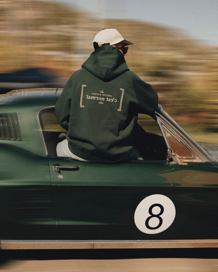 A man wearing Le Hoodie in Pine, a cap, and sunglasses, sits in the window of a moving dark green car. The image is a back view highlighting the French Riviera Club mirror logo with c'est normal text on the back of the hoodie.