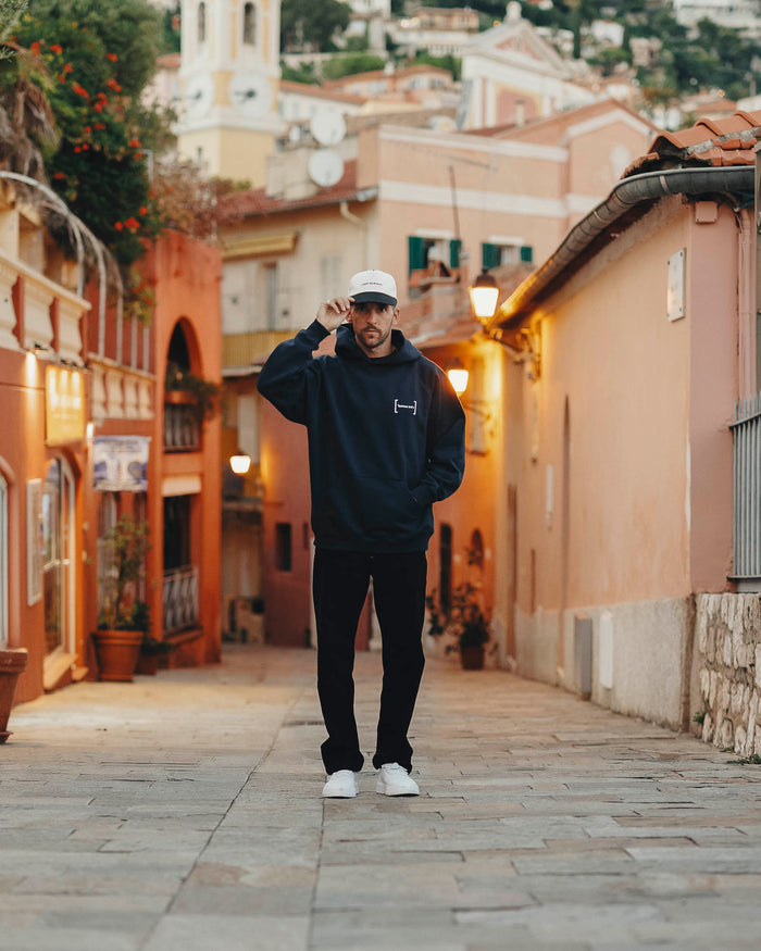 A man stands in a European town street wearing Le Hoodie in navy, an oversized hoodie featuring the white FRC mirror logo on the chest, paired with black pants, white sneakers, and a white c'est normal cap.