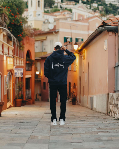 A back view of a man wearing Le Hoodie in navy, standing in a picturesque European alleyway. The image highlights the oversized fit and the white FRC mirror logo and c'est normal text on the center back.