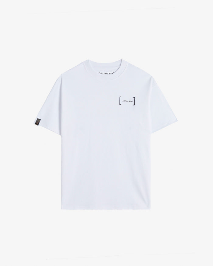 A front view of Le T-Shirt in white, laid flat on a white background. The image highlights the classic ribbed collar, the black FRC Edition mirror logo on the chest, and the signature black satin cuff tag with gold print from c'est normal.