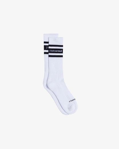 A pair of Les Socks in white, laid flat on a white background. The image highlights the mid-calf length, black contrast stitching with the c'est normal logo, and two black stripes.