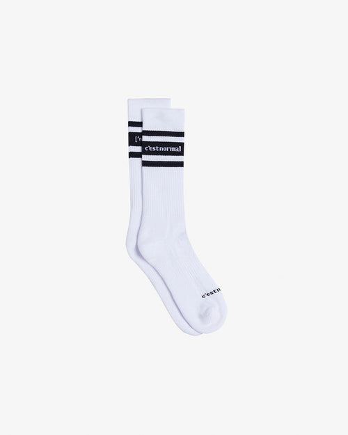 A pair of Les Socks in white, laid flat on a white background. The image highlights the mid-calf length, black contrast stitching with the c'est normal logo, and two black stripes.