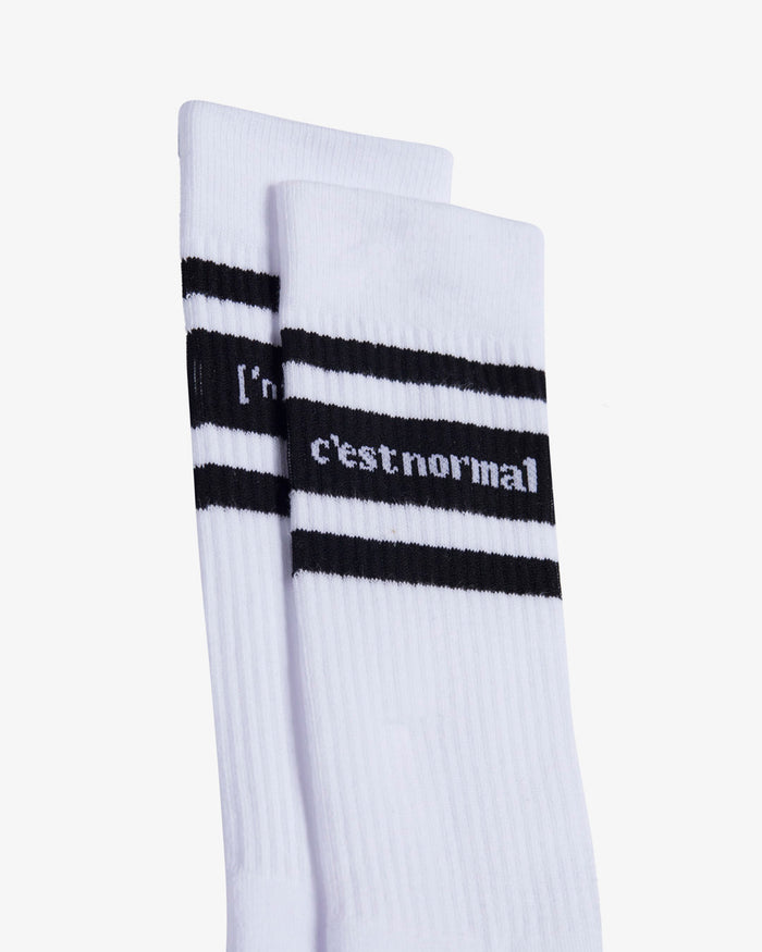 A close-up view of the upper part of Les Socks in white, against a white background. The image highlights the ribbed texture, the black horizontal stripes, and the c'est normal logo stitched mid-calf.