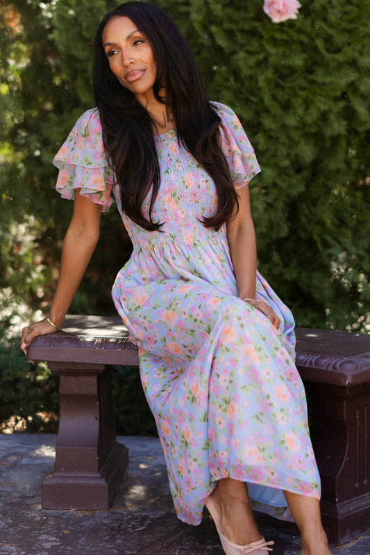 A woman with long dark hair sits on a brown bench outdoors, wearing the Ivy City Co Lottie Dress in Vibrant English Garden—a floral, maternity-friendly dress with ruffled sleeves—surrounded by greenery and sunlight.