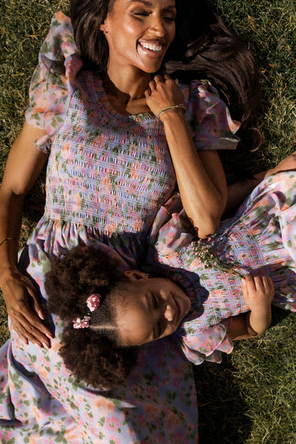 A woman and young girl lie on grass, smiling in matching Ivy City Co Lottie Dresses in Vibrant English Garden. The girl's hair is in puffs with pink clips. Sunlight highlights their joy in these maternity friendly dresses.