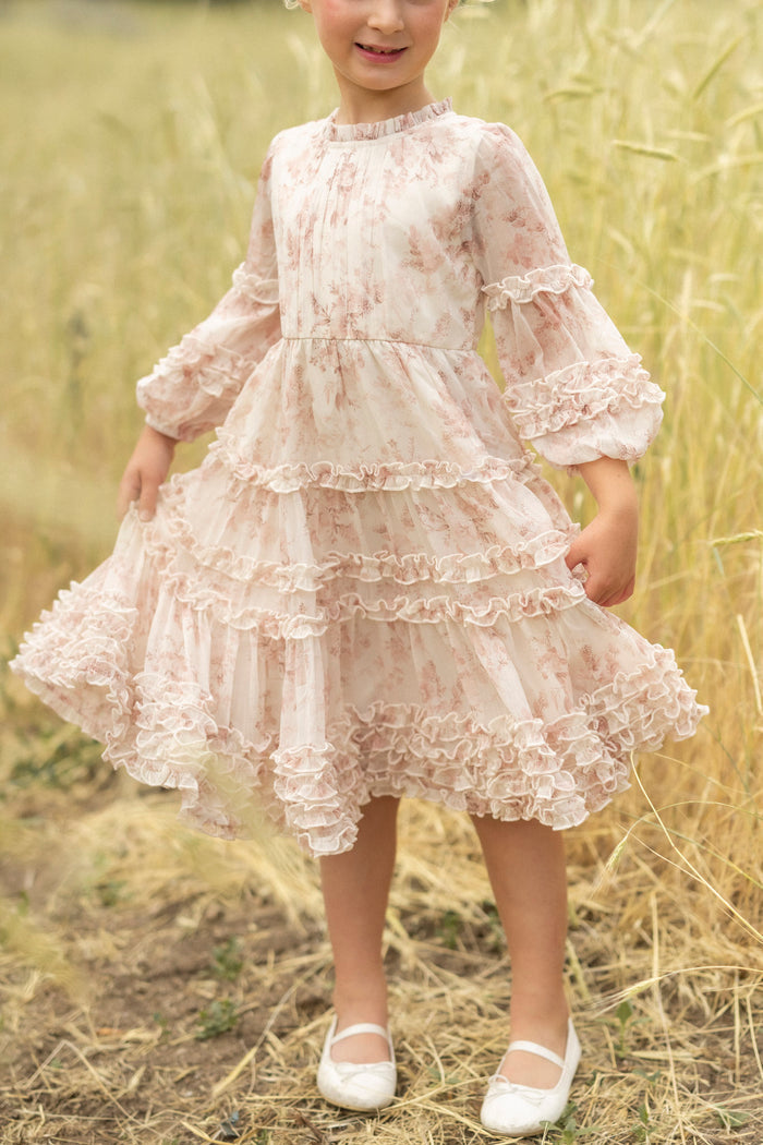 A young child stands in a grassy field, smiling and holding out the sides of her Ivy City Co Mini Geneva Dress in Banana Creme Aquarelle Fleur, a long-sleeved, ruffled floral dress. She pairs it with white shoes.