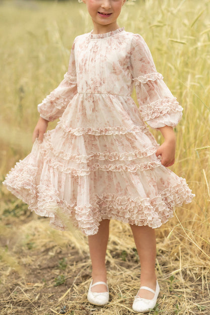 A young child stands in a grassy field, smiling and holding out the sides of her Ivy City Co Mini Geneva Dress in Banana Creme Aquarelle Fleur, a long-sleeved, ruffled floral dress. She pairs it with white shoes.