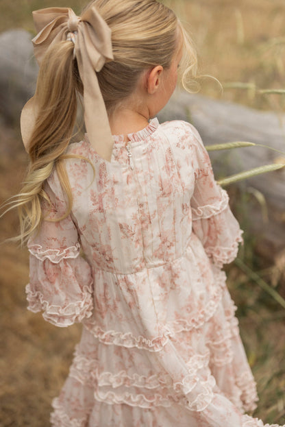 A young girl with blonde hair in a large beige bow wears the Ivy City Co Mini Geneva Dress in Banana Creme Aquarelle Fleur. She stands outdoors, back to the camera, near a wooden log and tall grass—perfect for a dreamy flower girl look.