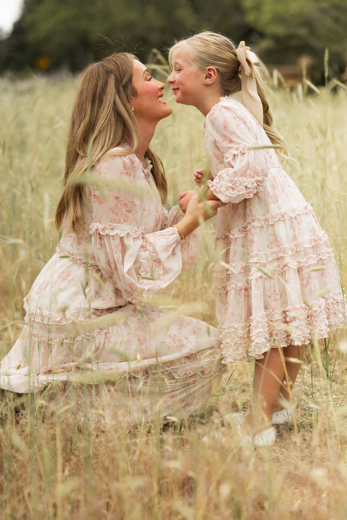 A woman kneels in a grassy field, smiling at a young girl. Both wear matching Ivy City Co Mini Geneva Dresses in Banana Creme Aquarelle Fleur, and the girl dons a big bow. They hold hands and share a joyful moment together.