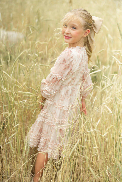 A young girl with a ponytail, wearing the Ivy City Co Mini Geneva Dress in Banana Creme Aquarelle Fleur and a large bow, stands in tall golden grass, looking over her shoulder and softly smiling at the camera.