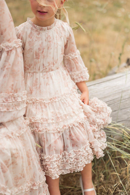 A young girl wears Ivy City Co's Mini Geneva Dress in Banana Creme Aquarelle Fleur outdoors by a fallen log, holding her yellow floral dress. Her face is partly visible, and she pairs the ruffled dress with white sandals.