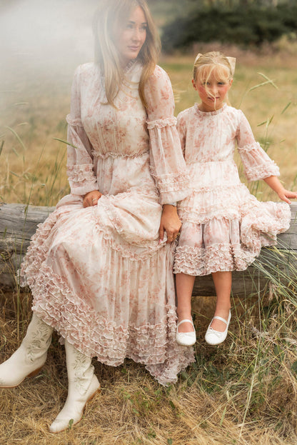 A woman and a young girl sit on a wooden log in a grassy field, both in Ivy City Co’s Mini Geneva Dress in Banana Creme Aquarelle Fleur. The girl pairs her dress with white shoes and cat-eye sunglasses.