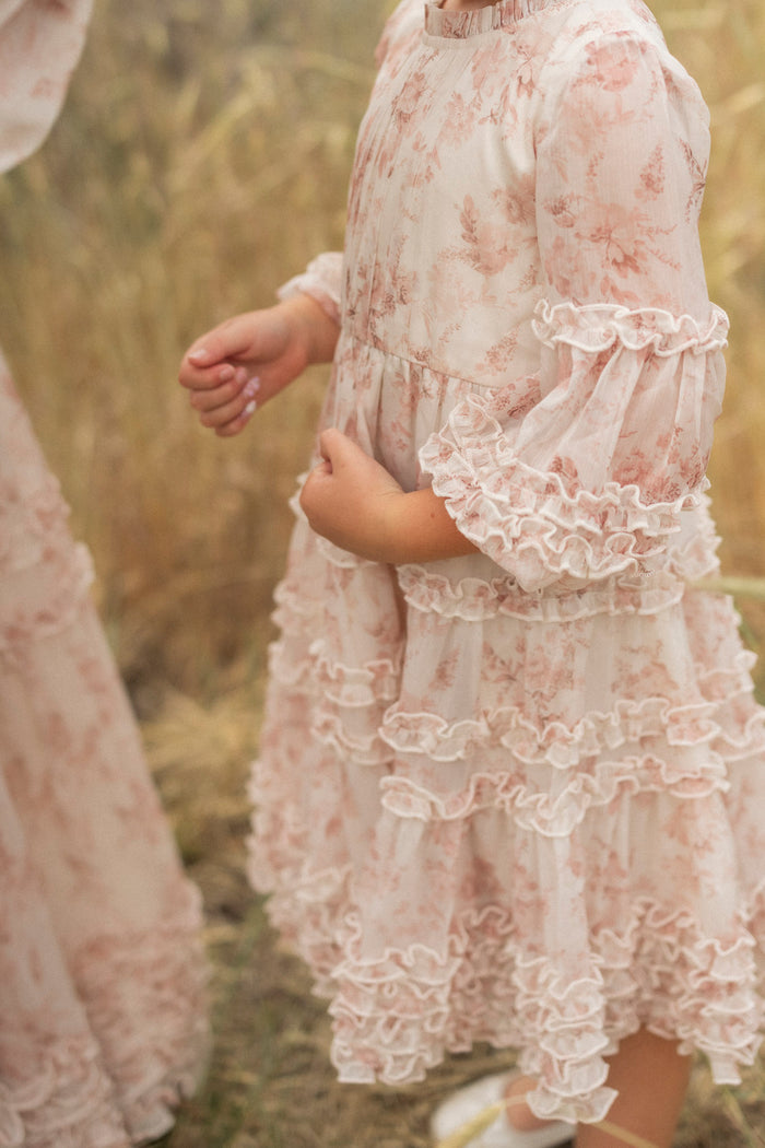 A young girl in the Ivy City Co Mini Geneva Dress in Banana Creme Aquarelle Fleur stands outdoors on dry grass, hands gently clasped. Only her midsection and arms are shown; another person in a similar flower girl dress is partly visible.