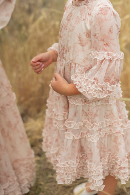 A young girl in the Ivy City Co Mini Geneva Dress in Banana Creme Aquarelle Fleur stands outdoors on dry grass, hands gently clasped. Only her midsection and arms are shown; another person in a similar flower girl dress is partly visible.