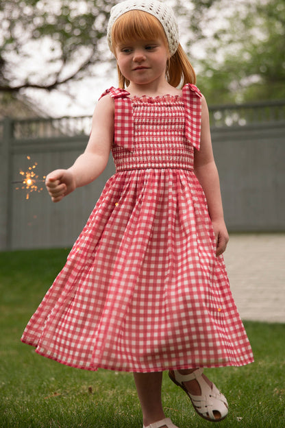 A young girl wears the Ivy City Co Mini Rayna Dress in Red Gingham and a white headscarf, holding a lit sparkler on grass with a gray fence and trees in the background.