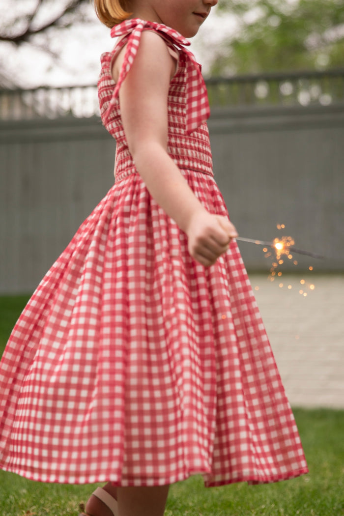 A young child wears the Ivy City Co Mini Rayna Dress in Red Gingham, holding a lit sparkler on grassy ground—perfect for a Mom and Me matching Fourth of July dress moment.
