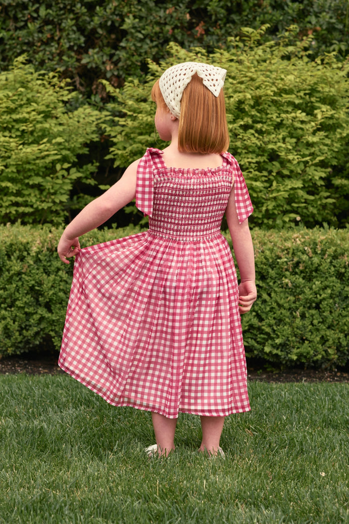 A young child with red hair and a white bow headband stands on grass, holding out one side of her Ivy City Co Mini Rayna Dress in Red Gingham. Green bushes and hedges are visible in the background.