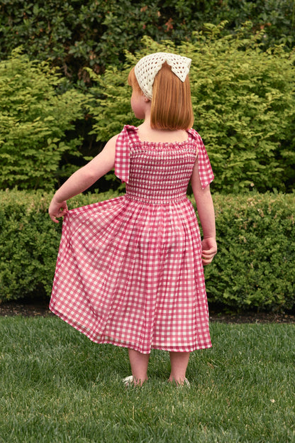 A young child with red hair and a white bow headband stands on grass, holding out one side of her Ivy City Co Mini Rayna Dress in Red Gingham. Green bushes and hedges are visible in the background.