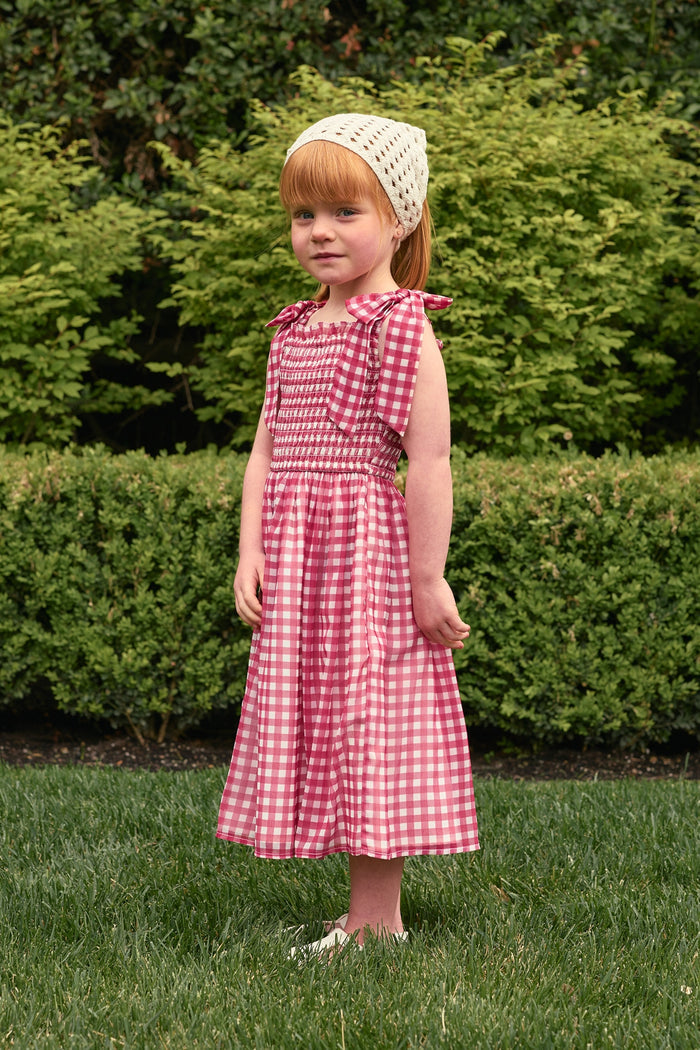 A young girl stands on grass in the Ivy City Co Mini Rayna Dress in Red Gingham, paired with white shoes and a crocheted headscarf, with green bushes behind her.