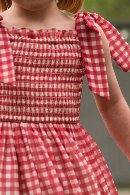 A child wears the Ivy City Co Mini Rayna Dress in Red Gingham with ruffled, bow-tied straps. The cropped photo highlights the dress and arms—an ideal classic look for the Fourth of July.