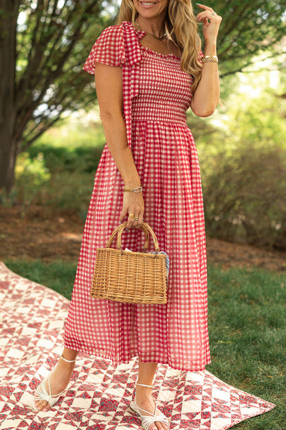 A woman wearing the Ivy City Co Rayna Dress in Red Gingham stands outside on a picnic blanket, holding a woven straw handbag. She's smiling with one hand in her hair and wears white sandals, surrounded by trees and greenery.