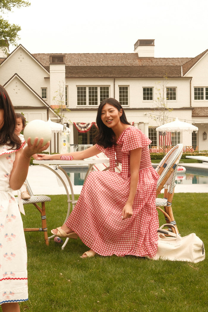 A woman smiles by a backyard pool wearing the Ivy City Co Rayna Dress in Red Gingham. Nearby, a child in a white dress with red trim holds a ball, with a large house visible in the background.