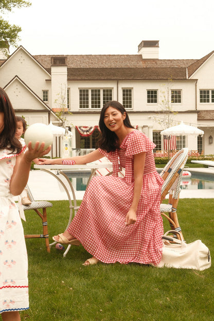 A woman smiles by a backyard pool wearing the Ivy City Co Rayna Dress in Red Gingham. Nearby, a child in a white dress with red trim holds a ball, with a large house visible in the background.