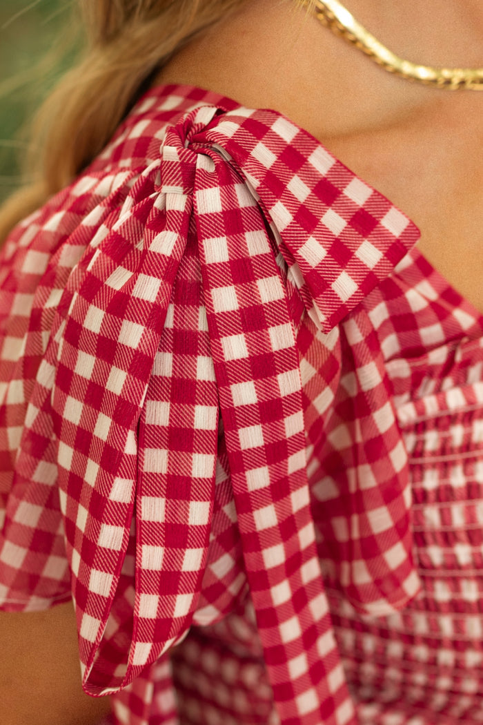Close-up of a person wearing the Rayna Dress in Red Gingham by Ivy City Co, featuring a smocked bodice, shoulder bow detail, and classic checkered pattern. A gold chain necklace completes the elegant look.