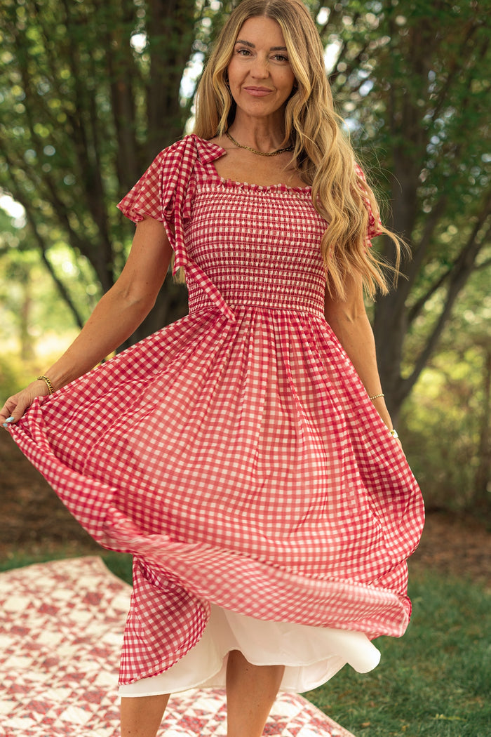 A woman stands outdoors on a checkered blanket, smiling as she holds the skirt of her Ivy City Co Rayna Dress in Red Gingham—a midi dress with short sleeves and a smocked bodice—surrounded by sunlight and green trees.