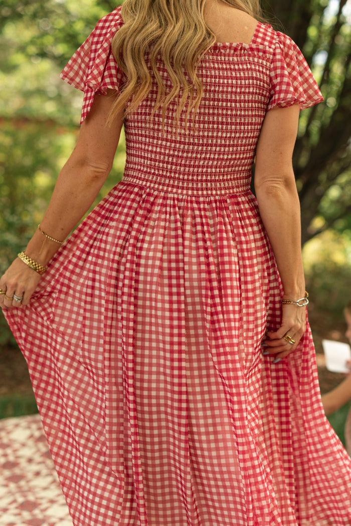 A woman with wavy blonde hair wears the Ivy City Co Rayna Dress in Red Gingham, featuring short flutter sleeves and a smocked bodice, holding the skirt out while standing outdoors amid a green, leafy background. She accessorizes with gold jewelry.