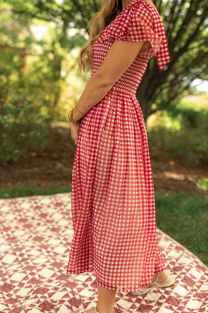 A woman stands outdoors on a patterned blanket, wearing the Ivy City Co Rayna Dress in Red Gingham—a midi dress with a smocked bodice and short sleeves. She has long, wavy hair and gold bracelets, surrounded by greenery and trees.
